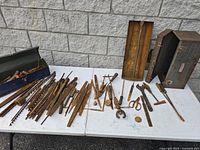 Wide view of old rusty hand tools including files, auger bits, and two metal toolboxes on white table surface against stone wall.