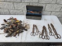 Overview photo showing the metal toolbox, four large wire cutters/tin snips, rusty scissors, and numerous small hand tools all on a white table.