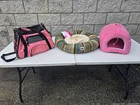 Photo of placed pink and black pet carrier, round striped pet bed, and pink cave bed on white table outside.