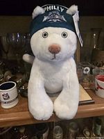 Front view of white plush animal wearing a green hat with Philly and WrestleMania 40 logos, sitting on a wooden surface with mugs in the background.