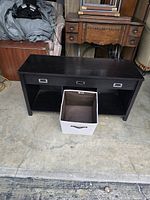Front view of small black wooden table with fabric storage container in front, showing table's flat top, open lower shelf, and two cut-out handles.