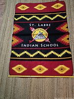 Blanket fully spread on floor showing central logo and tribal patterns in red, yellow, black with white text.