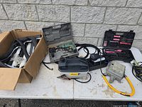 Photo showing sump pump, various drill and hand tool cases, spray paint containers, and pipe covers laid out on a white surface against an outdoor wall.