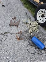 Three boat anchors displayed on pavement near trailer tire; includes two rusty iron anchors and one modern metal anchor with rope and blue buoy float.