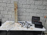 Two black tool boxes partially open showing various metal sockets, two white paint roller/brush strainers on white table with long rusty Rockwell metal part behind.