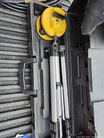 Folded tripod in a black carrying case showing aluminum legs and attached yellow leveling device with bubble level.