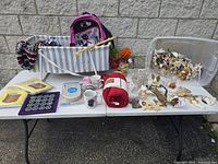 Photo showing vintage angel figurines, baby cups, red faux suede throw, striped backpack, and assorted household items on two tables against stone wall