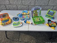Full view of six vintage battery operated educational toys arranged on white folding table outdoors.