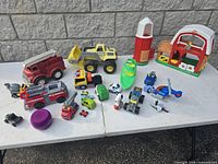 Full lot image showing farmhouse, silo, large dump truck, fire trucks, boat, helicopter, various small vehicles and figures on white table against stone wall.
