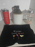 Photo showing three main bar accessory items: red metal traveling music box decanter, ceramic whiskey jug, and glass mixer with drink recipes. Black towel with embroidered drinking quote placed in front.