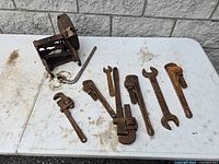 Photo shows eight rusty vintage pipe wrenches of various sizes and a rusty partially disassembled lawn mower part without handle on a white table against a gray brick wall.