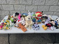 Lot of assorted vintage stuffed animals including Mario Bros characters, Mickey Mouse, and others displayed on white table outdoors against stone wall.