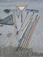 Seven yard tools including metal and plastic rakes, pitchfork, and wooden poles, laid out on gravel. Condition is weathered, used.