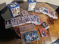 Overview of loose sports trading cards laid out on a table including football, basketball, and baseball players with some cards in a plastic bag and two sealed boxes seen in background.
