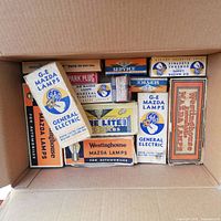 Top-down view of a cardboard box filled with various vintage automobile bulb boxes including multiple Westinghouse Mazda Lamps and General Electric G-E Mazda Lamps boxes.
