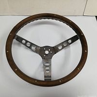 Circular antique steering wheel showing wooden rim, metal spokes with circular cutouts, and signs of wear.