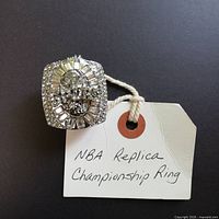 Front view of silver-tone replica NBA championship ring with rectangular face, embedded clear stones, and detailed sports design, placed next to handwritten tag "NBA Replica Championship Ring".