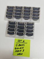 Overhead photo displaying 7 packs of Gillette Mach 3 razor blade refills, total 28 cartridges in clear plastic cases, arranged in two rows on a white surface with a yellow note identifying Set 2 and quantity.