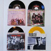 Four 45 RPM records with original picture sleeves: Bon Jovi, Sting, Parachute Club, John Cougar Mellencamp laid flat showing sleeves and vinyl discs.
