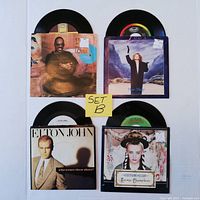 Four 45 RPM vinyl records with picture sleeves displayed flat, arranged in two rows. Top left is Stevie Wonder Part-Time Lover, top right Luba single. Bottom left Elton John, bottom right Culture Club Karma Chameleon.