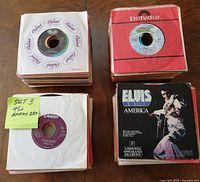 Two stacks of approximately 220 7-inch vinyl 45 RPM records in paper sleeves, some visible labels and sleeves, including Capitol Records and Polydor. One sleeve shows an Elvis Presley record titled 'America'.