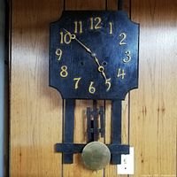 Front view of vintage black wooden Mission-style wall clock with gold numbers and brass pendulum visible below.