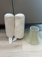 Photo showing two cylindrical white bolster cushion inserts with labels attached, next to a Pottery Barn lamp shade with a green checked fabric pattern.
