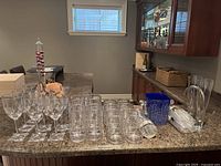 Photo of assorted clear acrylic and plastic glasses, including stemmed wine glasses, cylindrical beer glasses, blue acrylic ice holder, and many shot glasses on a kitchen countertop.