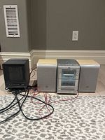 Photo showing a black ceramic heater on the left, silver mini audio system in the center with CD, tuner, and tape player, and two speakers on either side. All items are sitting on a carpeted floor against a wall.