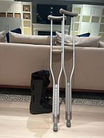 Pair of aluminum crutches standing upright next to a black walking boot on the floor in a living room setting.