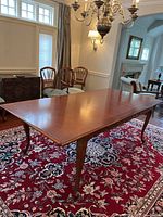 Maple dining table with single leaf in place shown on red patterned rug under chandelier