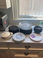 Photo showing multiple stacks of plates and bowls on a kitchen counter. Includes black Pfaltzgraff plates and bowls, blue bird design white plates, floral plates, and a white ceramic brie baking dish with lid.
