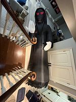 Full length view of large black inflatable Grim Reaper with red eyes and white-gloved hands suspended indoors, showing height and condition.