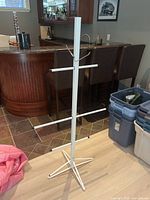 White metal hockey equipment drying rack with six horizontal arms on a vertical pole, supported by a three-legged base, standing indoors near a bar area.
