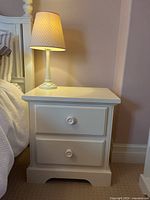 Front view of white painted wood nightstand with two drawers and round knobs, lamp on top