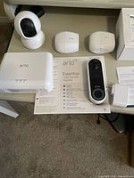 Set of Arlo security devices laid on carpet surface including two white Arlo wireless cameras, Netgear Arlo Base Station, two small white rectangular Arlo modules, wired power adapter, and Arlo Essential Video Doorbell box and paperwork.