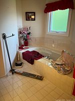 Overall view of bathroom corner showing tub with burgundy towel, decorative vase with flowers, Queen Elizabeth framed print on wall, two black curtain rods on floor, a bathroom scale, and shower curtain.