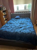 Full view of double bed set up with blue linens, grey headboard and frame, next to window with floral wallpaper and red curtains.