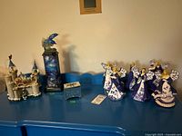 Photo showing full lot on blue table including six Blue Willow angel figurines, dolphin pillar figurine, castle with Christmas snow globes, and blue mirrored trinket box.