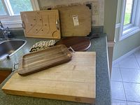 Five different wood boards and one kitchen apron arranged on kitchen counter around sink and windows. Boards include a large butcher block style board, turkey carving board, drawer-style with handles, and paddle-shaped board.