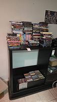 Large collection of DVDs and CDs stacked on a black media cabinet. DVDs include movies and series, many titles stacked vertically and horizontally.