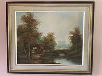 Full framed painting of a cottage landscape with a waterwheel by a river and stone bridge, surrounded by trees with autumnal colors.