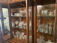 Three shelves of assorted glassware, including wine glasses, crystal decanter, candy dishes, and more, displayed inside a wooden cabinet.