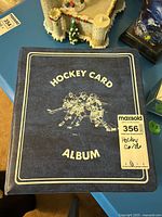 Blue Hockey Card Album binder with stamped text 'Hockey Card Album' and drawing of three hockey players skating in formation on the front cover.