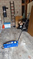 Blue Michelin hydraulic jack with handle extended on garage floor with ladders and garage items in background.