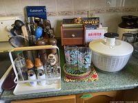 Overview photo showing spice rack with shakers, cookbooks, two drinking glasses on crocheted coaster, wooden recipe box, salad spinner, and utensils.