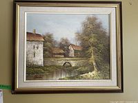 Full view of the framed landscape painting showing a river, stone bridge, and rustic buildings under trees.