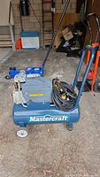 Blue Mastercraft wheeled air compressor with coiled black hose and attached spray gun, with visible Mastercraft label and handle.