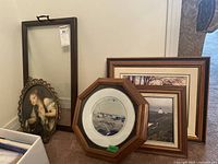 Lot of framed vintage art prints, framed photos, collector plate and serving tray arranged against a wall.