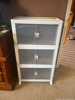 White plastic shelving unit with three cabinet-style compartments closed, showing ribbed semi-transparent fronts and silver handles, standing on four caster wheels.